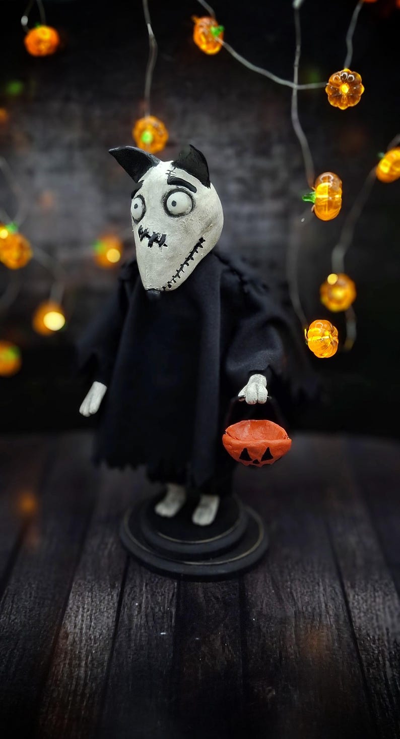 Handmade Sparky Art Doll wearing a black hoodie Halloween-style outfit, holding a pumpkin-shaped trick-or-treat box. Shown from a right diagonal angle, looking downward. Whimsical and spooky Halloween home decor, unique collectible dog doll