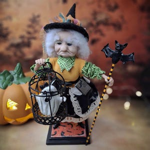 Front view of a handmade witch art doll turned slightly to the left, wearing a Halloween-themed outfit and holding a cage with a tiny ghost in one hand and a bat-shaped wand in the other, looking at the cage.