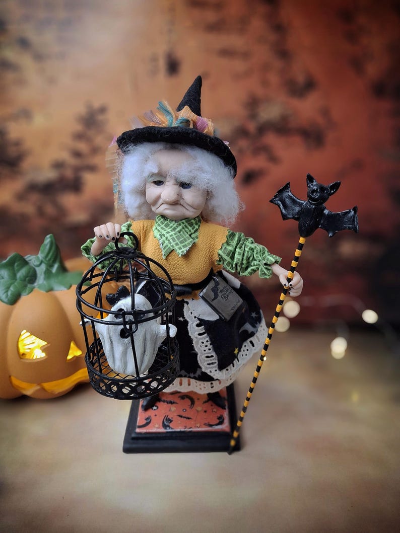 Front view of a handmade witch art doll turned slightly to the left, wearing a Halloween-themed outfit and holding a cage with a tiny ghost in one hand and a bat-shaped wand in the other, looking at the cage.