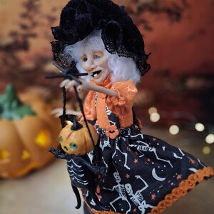 Side view of the handmade witch art doll from the left, showing her Halloween-themed outfit, detailed hand-painted face, and the tiny pumpkin skeleton puppet she holds.