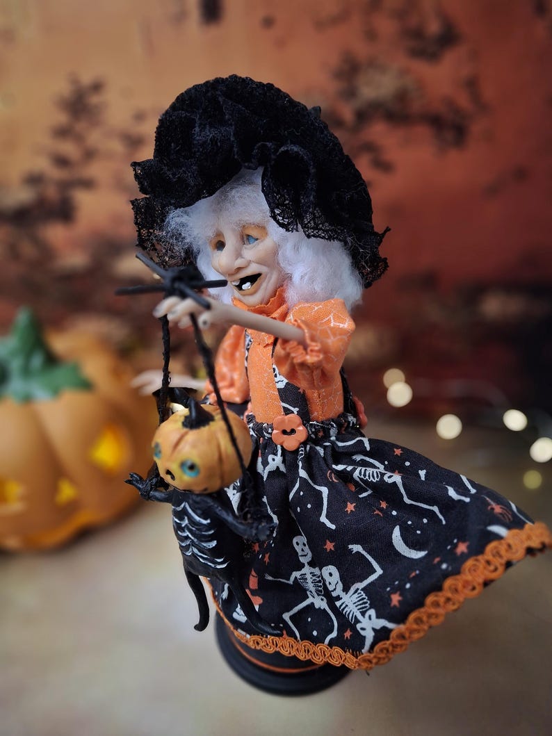 Side view of the handmade witch art doll from the left, showing her Halloween-themed outfit, detailed hand-painted face, and the tiny pumpkin skeleton puppet she holds.