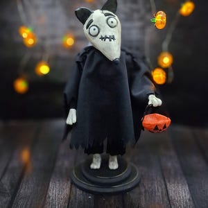Wide shot of Handmade Sparky Art Doll wearing a black hoodie Halloween-style outfit, holding a pumpkin-shaped trick-or-treat box. Standing straight, facing slightly to the right. Unique whimsical and spooky Halloween home decor collectible dog doll