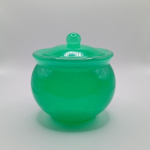 Könnte beinhalten: Ein leuchtend grünes Glasgefäß mit passendem Deckel. Das Gefäß hat eine runde Form und einen kleinen Knopf auf dem Deckel. Das Glas hat ein leicht gesprenkeltes Aussehen, und das Gefäß steht vor einem weißen Hintergrund.