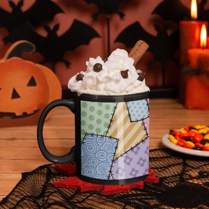 May include: Black coffee mug with a patchwork design featuring different colors and patterns. The mug is filled with whipped cream and chocolate chips, and a cinnamon stick is sticking out of the top. The mug is sitting on a black lace doily with a red felt coaster.
