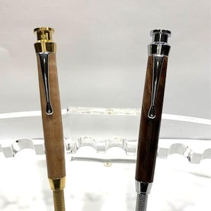 May include: Two wooden pens with metal accents. The pen on the left has a gold top and the pen on the right has a silver top. Both pens are sitting in a clear acrylic stand.