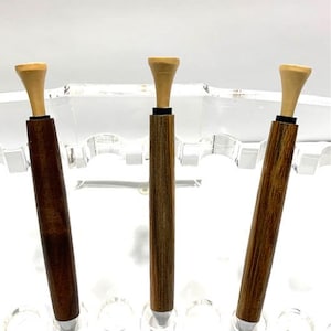 May include: Three wooden golf ball markers with a beige plastic golf tee on top. The markers are different shades of brown wood.