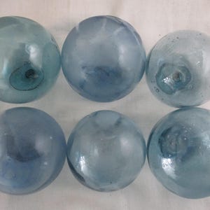 May include: Six spherical glass fishing floats in varying shades of light blue and translucent gray. Each orb features a distinct, slightly irregular surface texture with small air bubbles and a central sealed glass pontil mark on one side.