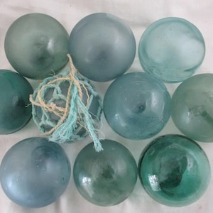 May include: A collection of nine glass fishing floats in shades of blue and green. One float is encased in a blue net with a rope tie. The floats vary in size and color, creating a coastal aesthetic.