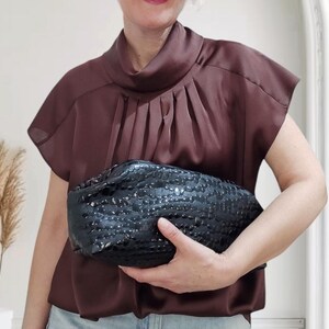 Könnte beinhalten: Eine schwarze Clutch mit einem strukturierten, gewebten Design wird vor einer Person gehalten. Die Clutch ist der Mittelpunkt, mit einer dunkelbraunen Bluse und hellblauen Jeans im Hintergrund. Die Bluse hat einen hohen Ausschnitt und Falten.