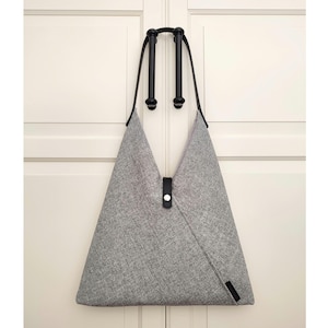 May include: A gray triangular tote bag with black leather straps and accents. The bag features a button closure and a small black tag. The bag is hanging on a white door.