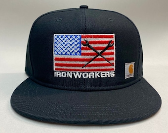 May include: Black Carhartt fitted baseball cap with an American flag and crossed wrenches embroidered on the front. The text "IRONWORKERS" is embroidered below the flag. The Carhartt logo is on the right side of the cap.