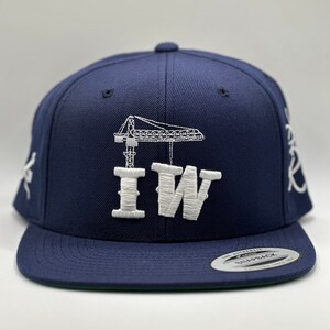 Peut inclure: Une casquette de baseball bleu marine avec une grue brodée en blanc et les lettres "IW". La casquette a une sangle réglable noire et une étiquette blanche "The Authentic Snapback".