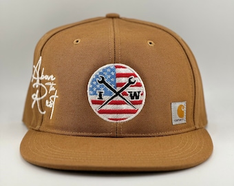 May include: A brown Carhartt brand baseball cap with a white embroidered American flag patch featuring crossed wrenches and the letters "I" and "W". The text "Above the Rest" is embroidered in white on the side of the cap.