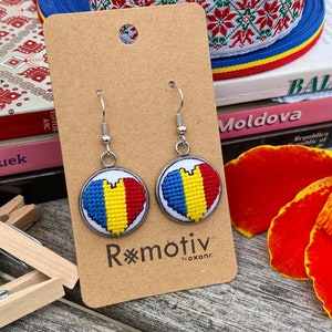 May include: A pair of heart-shaped earrings featuring a cross-stitch design of the Romanian flag. The earrings are silver and have a hook fastening. The earrings are displayed on a brown card with the text "R*motiv" and "boxanc".