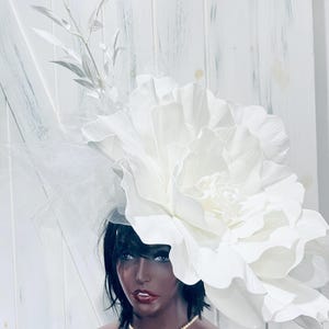 May include: A large, white floral headpiece with a layered petal design. The headpiece is accented with delicate white leaves and tulle. The mannequin is wearing a pearl necklace and has dark hair.