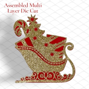 May include: An assembled multi-layer die cut featuring a gold glitter sleigh filled with a gold glitter Christmas tree, a candy cane, and red gift boxes. The design includes intricate scrollwork at the base and star accents on the tree.