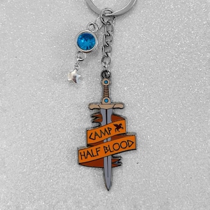 Op de afbeelding: Een zilverkleurige sleutelhanger met een zwaardhanger en een oranje banner met de tekst "CAMP HALF BLOOD". De sleutelhanger bevat ook een blauwe edelsteen en een kleine sterrenbedel.