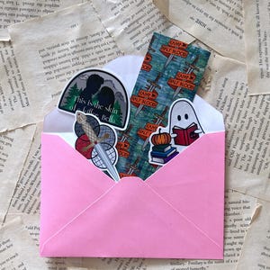 Puede incluir: Un sobre rosa que contiene pegatinas y un marcapáginas. Las pegatinas presentan texto e ilustraciones, incluyendo una silueta, una espada y un fantasma leyendo. El marcapáginas tiene un diseño de espada. Los artículos están sobre un fondo de páginas de libro.