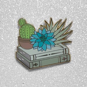 Pode incluir: Um broche de esmalte dourado com uma pilha de três livros com um cacto verde e uma suculenta azul no topo.