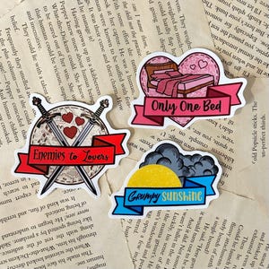 Op de afbeelding: Vier stickers op een pagina uit een boek. Eén zegt "Enemies to Lovers" met gekruiste zwaarden en harten. Een andere zegt "Only One Bed" met een bedgrafiek. De derde zegt "Grumpy Sunshine" met een zon en wolken.