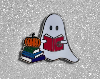 May include: A white ghost with black eyes is reading a red book with a skull on the cover. The ghost is holding a stack of books and a small orange pumpkin.