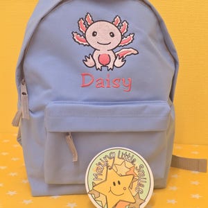 May include: A pale blue backpack with a front pocket and zip. It has an embroidered axolotl design in pink and white, with the name "Daisy" embroidered below. A round sticker with a smiling star is in front of the backpack.