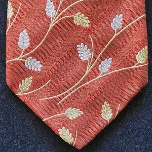 May include: A rust-colored necktie with a repeating pattern of embroidered leaves and branches in silver, gold, and cream. The tie has a textured appearance and a classic pointed end. This accessory is suitable for formal or business attire.
