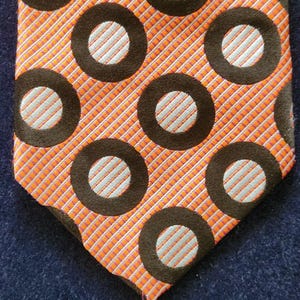 Puede incluir: Primer plano de una corbata estampada. La corbata presenta un patrón repetitivo de grandes círculos marrones con centros a rayas blancas y grises sobre un fondo naranja. La corbata está hecha de tela texturizada.