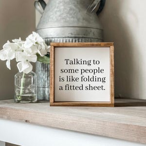 Peut inclure: Un panneau encadré en bois avec le texte "Talking to some people is like folding a fitted sheet." Le panneau est posé sur une surface en bois, avec une cruche en métal galvanisé et un vase de fleurs blanches en arrière-plan.