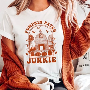 May include: White t-shirt with an orange graphic of a farmhouse, pumpkins, and the text "Pumpkin Patch Junkie".