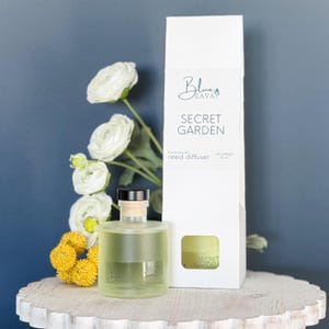 May include: A clear glass reed diffuser with a black cap and a white box. The box reads "Secret Garden" and "hand poured reed diffuser". The diffuser is next to white flowers and yellow flowers. The net weight is 12 oz.