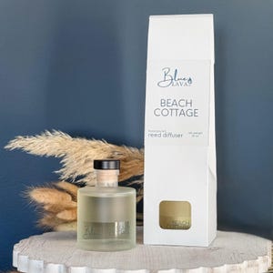 May include: A clear glass reed diffuser bottle with a black cap and a white box. The box and bottle both have the text "Beach Cottage" and the brand logo. The diffuser is next to a white box with the same branding.