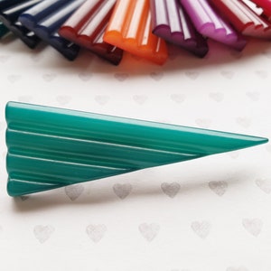 May include: A teal-colored, triangular hair clip with a ridged design. The clip is set against a white background with heart patterns, with a fan of colorful hair clips in the background. The other clips are in shades of purple, red, orange, and blue.