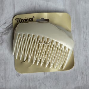Peut inclure: Un peigne à cheveux en plastique de couleur crème avec un bord supérieur incurvé et plusieurs dents, fixé sur un support en carton beige portant le nom de marque Riviera imprimé en noir avec une découpe pour suspension.