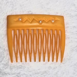 May include: An amber-coloured vintage hair comb. The top edge features a zigzag design with three small, pearl-like accents. The comb has long, evenly spaced teeth, a classic hair accessory. The comb's design suggests a retro style.
