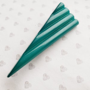 May include: A teal-colored, triangular-shaped object with a ridged top. The object has a pointed end and a series of parallel ridges along its top surface. The background is a white surface with a pattern of small gray hearts.