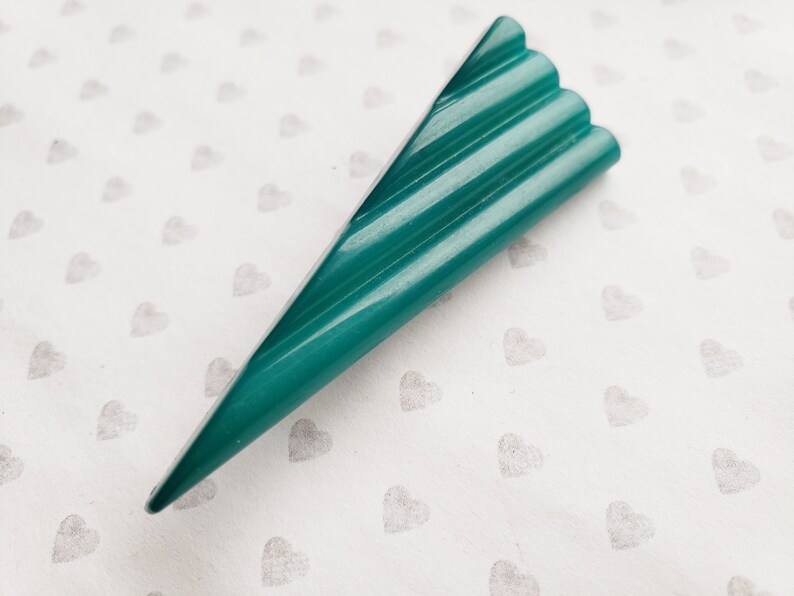 May include: A teal-colored, triangular-shaped object with a ridged top. The object has a pointed end and a series of parallel ridges along its top surface. The background is a white surface with a pattern of small gray hearts.
