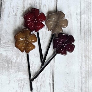 May include: Four flower-shaped hair pins in shades of red, brown, and tan. Each pin has a black stem. The flowers have a glossy finish and are arranged on a white wooden surface.