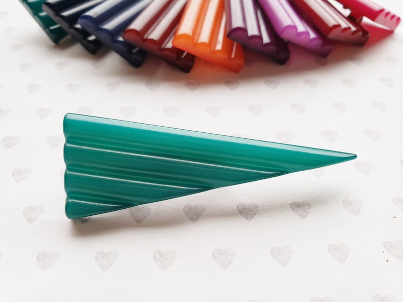 May include: A teal-colored, triangular hair clip with a ridged design. The clip is set against a white background with heart patterns, with a fan of colorful hair clips in the background. The other clips are in shades of purple, red, orange, and blue.