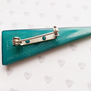 May include: Teal-colored triangular brooch with a silver pin backing. The brooch has the text "MODELE DEPOSE" and "BUCH + DEICHMANN DENMARK" printed on it. The brooch is a vintage accessory.