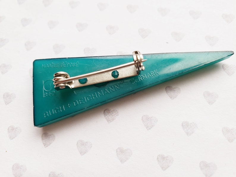 May include: Teal-colored triangular brooch with a silver pin backing. The brooch has the text "MODELE DEPOSE" and "BUCH + DEICHMANN DENMARK" printed on it. The brooch is a vintage accessory.