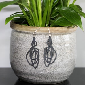 May include: A pair of black dangle earrings with a geometric design. The earrings are hanging from a white ceramic pot with a crackled glaze.