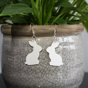 May include: A pair of white bunny silhouette earrings with silver hooks. The earrings are hanging from a potted plant.