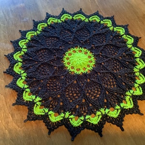 May include: A black and lime green crocheted doily with a circular design. The doily has a scalloped edge and a central motif.