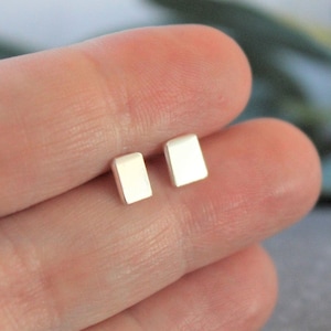 May include: A pair of small, rectangular silver stud earrings. The earrings have a slightly curved top and a polished finish. The earrings are displayed against a blurred background.