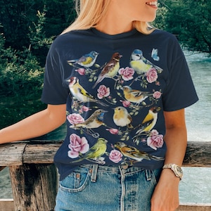 以下が含まれることがあります： ネイビーブルーのTシャツ。水彩画のデザインで、鳥とピンクのバラが描かれています。鳥は黄色、青、茶色、緑の色合いです。カジュアルなスタイルのTシャツです。