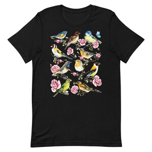 以下が含まれることがあります： 黒いTシャツ。水彩画のデザインで、ピンクのバラの枝に止まっている様々なカラフルな鳥が描かれています。鳥は青、黄色、茶色、緑の色合いです。小さな青い蝶もデザインに含まれています。