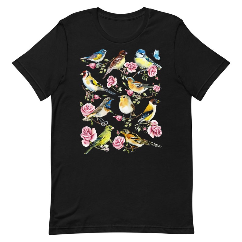以下が含まれることがあります： 黒いTシャツ。水彩画のデザインで、ピンクのバラの枝に止まっている様々なカラフルな鳥が描かれています。鳥は青、黄色、茶色、緑の色合いです。小さな青い蝶もデザインに含まれています。