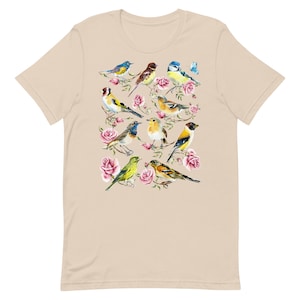 以下が含まれることがあります： ベージュ色のTシャツ。水彩画で描かれた、ピンクのバラの枝にとまる様々なカラフルな鳥のデザイン。鳥は青、黄色、茶色、緑色です。Tシャツは半袖で、クルーネックです。