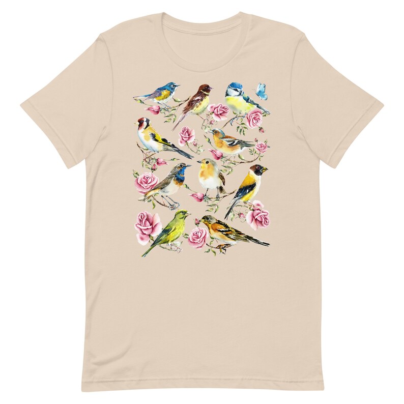 以下が含まれることがあります： ベージュ色のTシャツ。水彩画で描かれた、ピンクのバラの枝にとまる様々なカラフルな鳥のデザイン。鳥は青、黄色、茶色、緑色です。Tシャツは半袖で、クルーネックです。